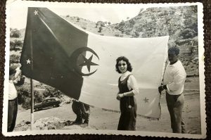 Photo prise le 19 mars 1962 à Oued Fodda par Zoubir Slamani représentant Baya Slamani devant un grand drapeau algérien et souriant. Le drapeau est porté par deux personnes et derrière lui, les pieds d'un maquisard en tenue militaire apparaissent (archives personnelles de Baya Slamani). 